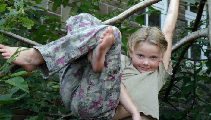 Young feral boy smiling while swinging fearlessly on a branch, wearing a light green shirt and camouflage pants.
