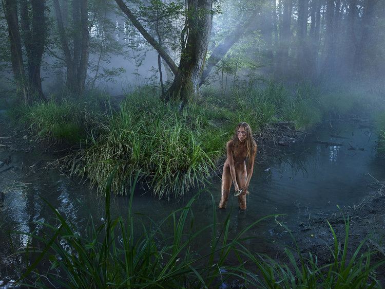 A topless feral woman in a swamp in the wild, with a serious face and blonde hair.