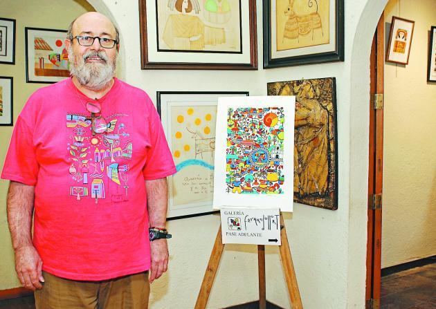 Fernando Llort smiling while standing beside his painting and wearing a pink printed t-shirt, eyeglasses, brown pants, and wristwatch