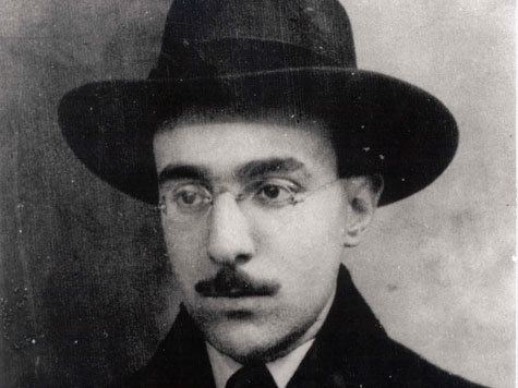 Fernando Pessoa is serious, in black and white, has black hair mustache wearing a black panama hat, eyeglasses, white polo and black necktie, under a black coat.