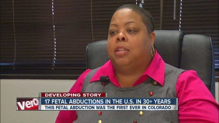On a news report, Allison Cotton has black short hair, below is a report about the developing story of “17 Fetal Abductions In The U.S. In 30+ Years, This Fetal Abduction Was The First Ever In Colorado” she is sitting down on a black chair, behind her are black blinds, she is wearing gold earrings, white-red necklace, a small microphone attached on her pink polo under a gray vest.