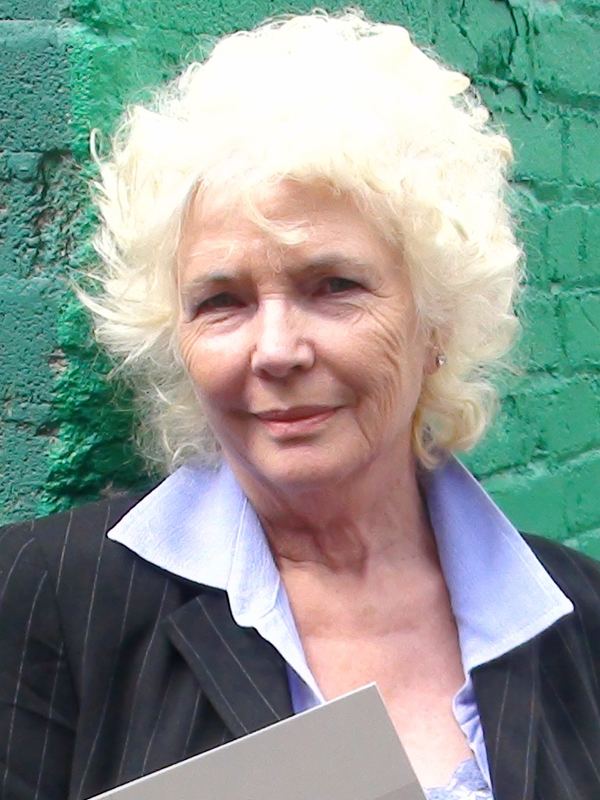 Fionnula Flanagan leaning on a wall and wearing a sky blue formal dress underneath a black blazer.