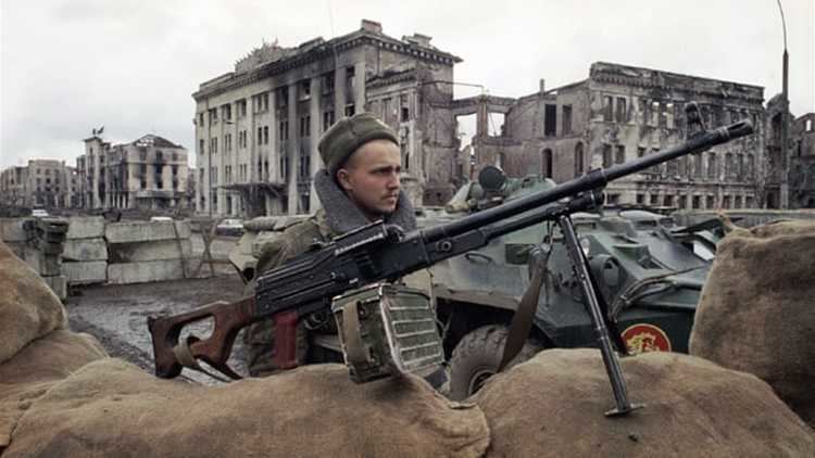 A Russian soldier with a large caliber rifle beside him during the Chechen War while destroyed buildings can be seen behind him