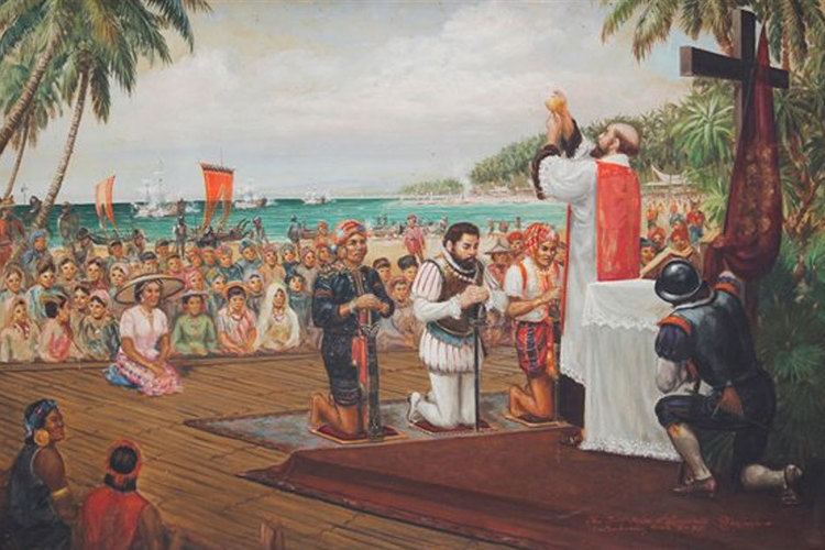 The painting of the First Mass in the Philippines, at the back is a shore with boats and flags on it, along with the crowd and citizen of two different counties, in front is a three men kneeling in front of a priest, from left, a man kneeling on a pillow while holding a sword with both hands wearing a red cloth and a black native clothing, 2nd from left is a european crew, Ferdinand Magellan is kneeling looking down holding his sword with his hands, has brown hair, mustache and beard, wearing a white clothing and brown vest, 3rd from left, a man is kneeling, with his head down, hands together holding a sword, wearing a red head cover a white shirt and a red cloth around his waist, a the right is the priest hands up, holding a chalice while standing in front of a white table with a cross, and a red flag holding by a european crew, has bald top and brown side hair mustache and beard, wearing a priest white long robe with red stole,