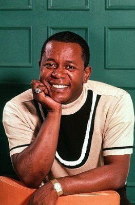 Flip Wilson is smiling while leaning on an orange chair and his right hand is on his face, wearing a ring, a watch, and a white and black shirt.
