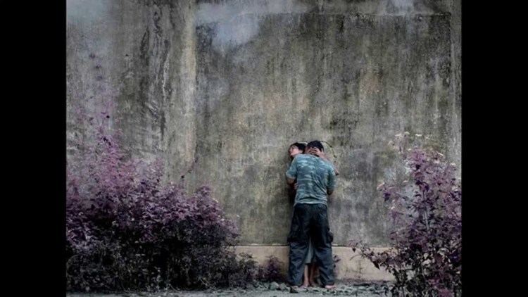 Chaminda Sampath Jayaweera kissing Gayesha Perera on the neck. Gayesha is leaning on the wall with purple plants on both sides and holding Chaminda's neck while her eyes are closed, wearing earrings in a scene from the 2011 anthology film, Flying Fish. Chaminda is wearing a green and blue camouflage t-shirt while Gayesha is wearing a gray dress.