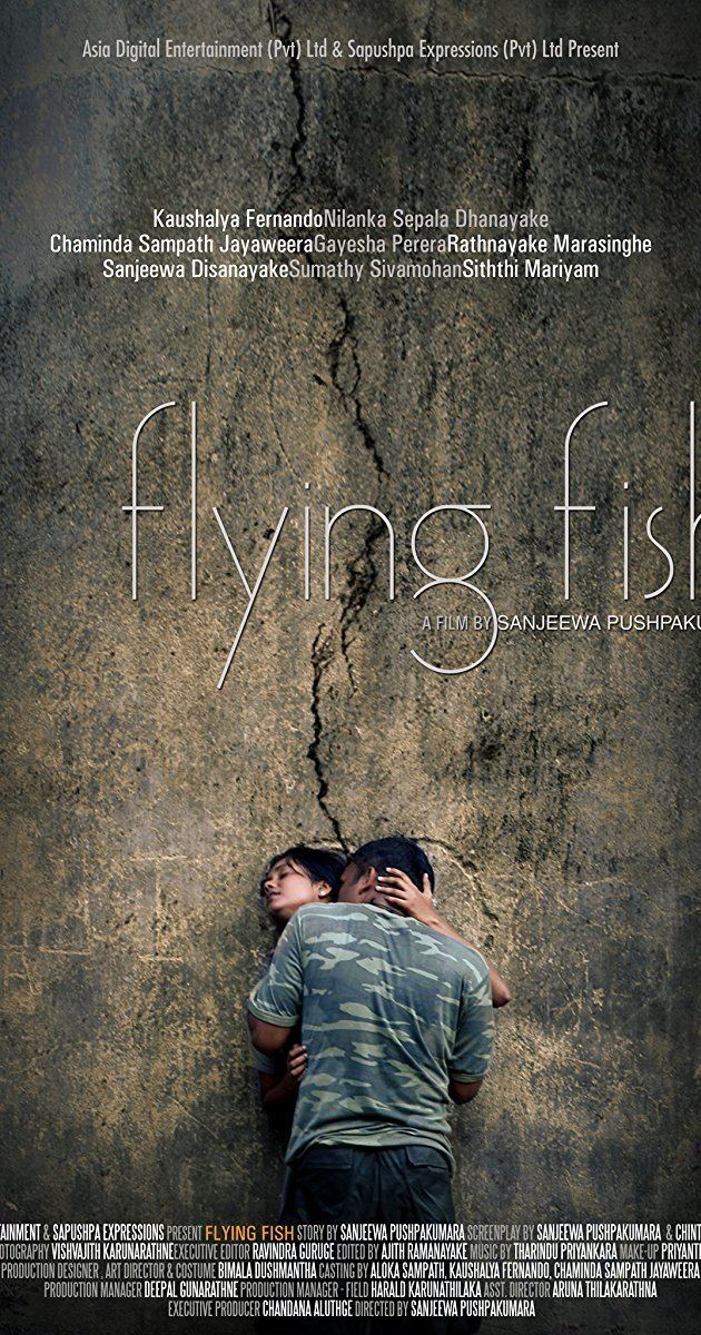 A movie poster of the 2011 film "Flying Fish" starring Gayesha Perera as Wasana and Chaminda Sampath Jayaweera as a soldier. Chaminda kissing Gayesha on the neck. Gayesha is leaning on the wall and holding Chaminda's neck while her eyes are closed. Chaminda is wearing a green and blue camouflage t-shirt while Gayesha wearing earrings and a gray dress