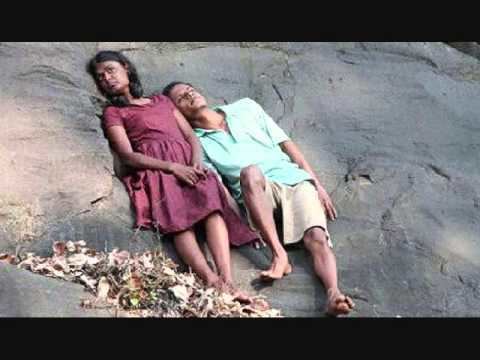 Gayesha Perera and Nilanka Dahanayake are looking afar while leaning on a big rock with serious faces and dried leaves on their feet in a scene from the 2011 anthology film, Flying Fish. Gayesha is wearing a pink dress while Nilanka is wearing a blue polo shirt and khaki shorts.