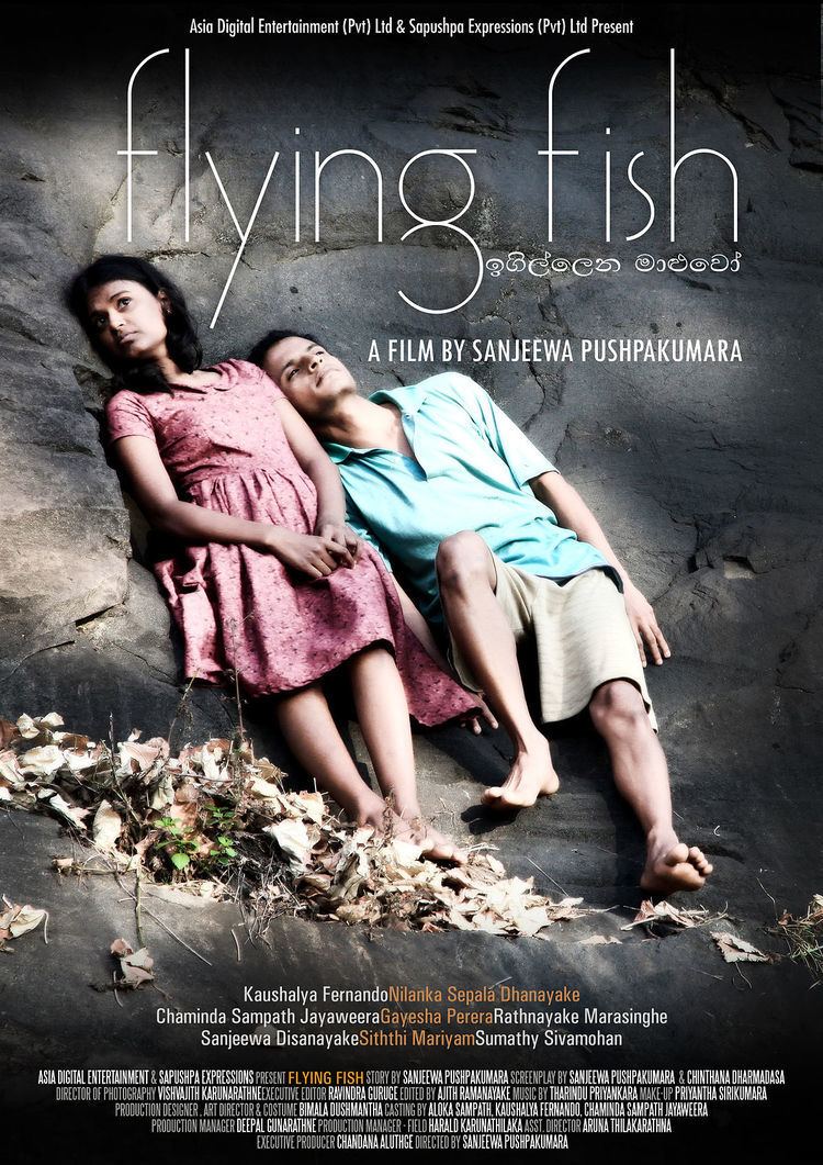 The movie poster of the 2011 anthology film "Flying Fish". Gayesha Perera and Nilanka Dahanayake are looking afar while leaning on a big rock with serious faces and dried leaves on their feet. Gayesha is wearing a pink dress while Nilanka is wearing a blue polo shirt and brown shorts