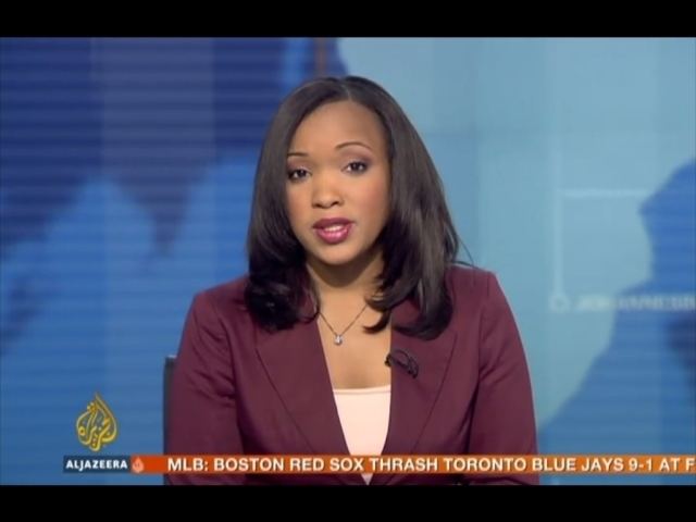 Folly Bah Thibault reporting the news while wearing a maroon coat, light pink inner blouse, and necklace