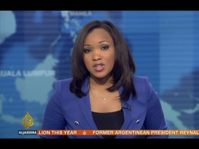 Folly Bah Thibault reporting the news while wearing a blue coat, black inner blouse, and necklace