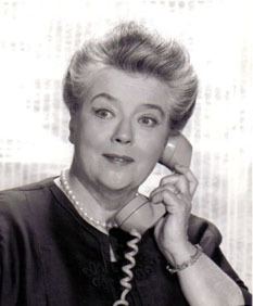 Frances Bavier with a tight-lipped smile while holding the telephone and wearing a blouse and pearl necklace