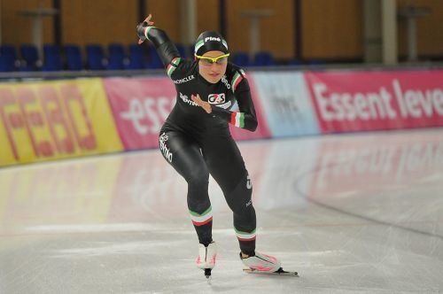 Francesca Lollobrigida doing speed skating, wearing yellow protective sports glasses, a multi-colored aerodynamic racing suit with a hood, and white and pink skates.