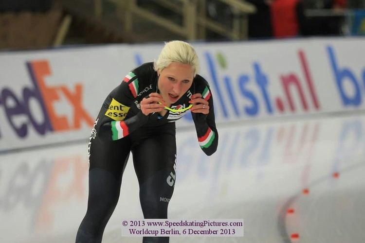 Francesca Lollobrigida panting while doing speed skating and holding yellow protective sports glasses, with blonde hair, and wearing a multi-colored aerodynamic racing suit.