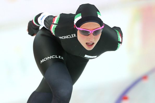 Francesca Lollobrigida with a determined face while doing speed skating, wearing purple protective sports glasses, and a multi-colored aerodynamic racing suit with a hood.