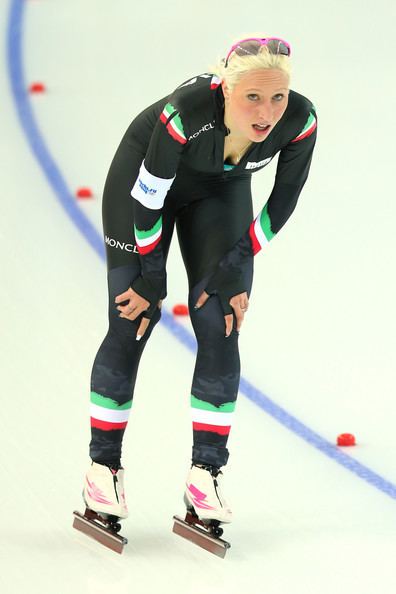 Francesca Lollobrigida panting while doing speed skating, with blonde hair, wearing purple protective sports glasses, a multi-colored aerodynamic racing suit, and white and pink skates.