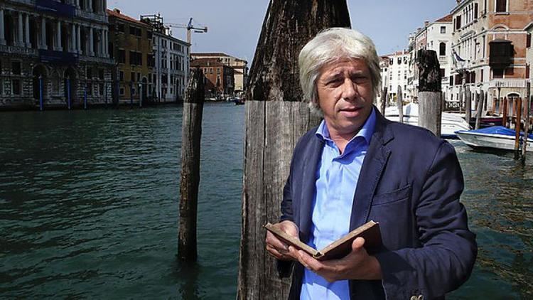 Francesco da Mosto smiling and holding a book while wearing black coat and blue long sleeves