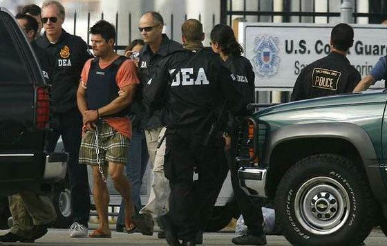 Francisco Javier Arellano Felix with a handcuffed while surrounded by the Coast Guardsmen, wearing a blue vest, orange shirt, brown checkered shorts, and slippers.