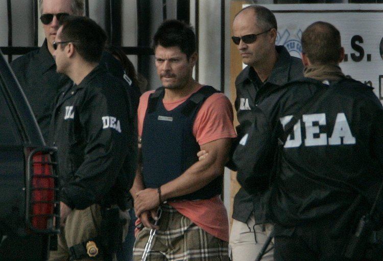 Francisco Javier Arellano Felix with a handcuffed while surrounded by the Coast Guardsmen, wearing a blue vest, orange shirt, and brown checkered shorts.