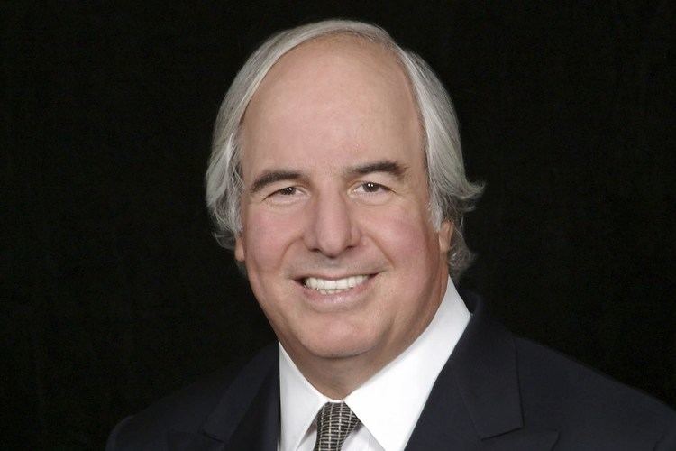 Frank Abagnale smiling and wearing a black coat over a white suit and a necktie.