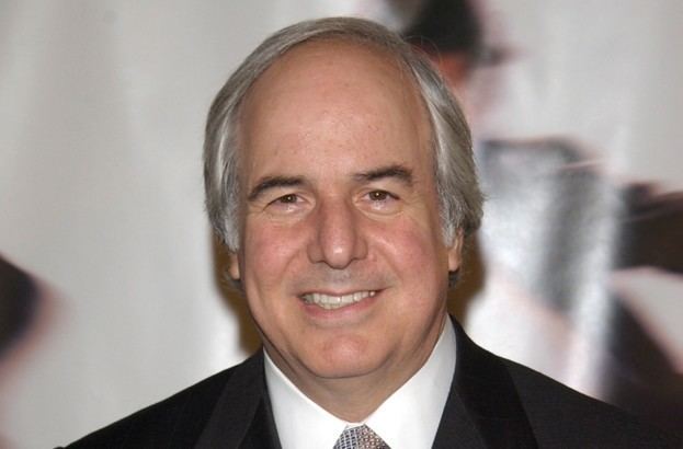 Frank Abagnale smiling and wearing a black coat over a white suit and a necktie.