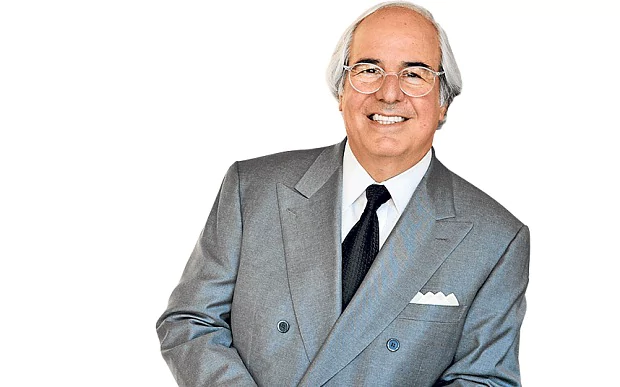 Frank Abagnale smiling and wearing a graycoat over a white suit and a black necktie as well as an eyeglass.
