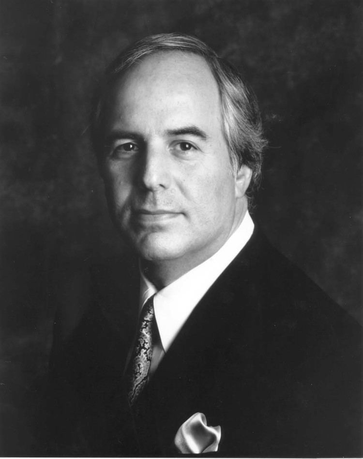 A younger Frank Abagnale posing and wearing a black coat over a white suit and a necktie.