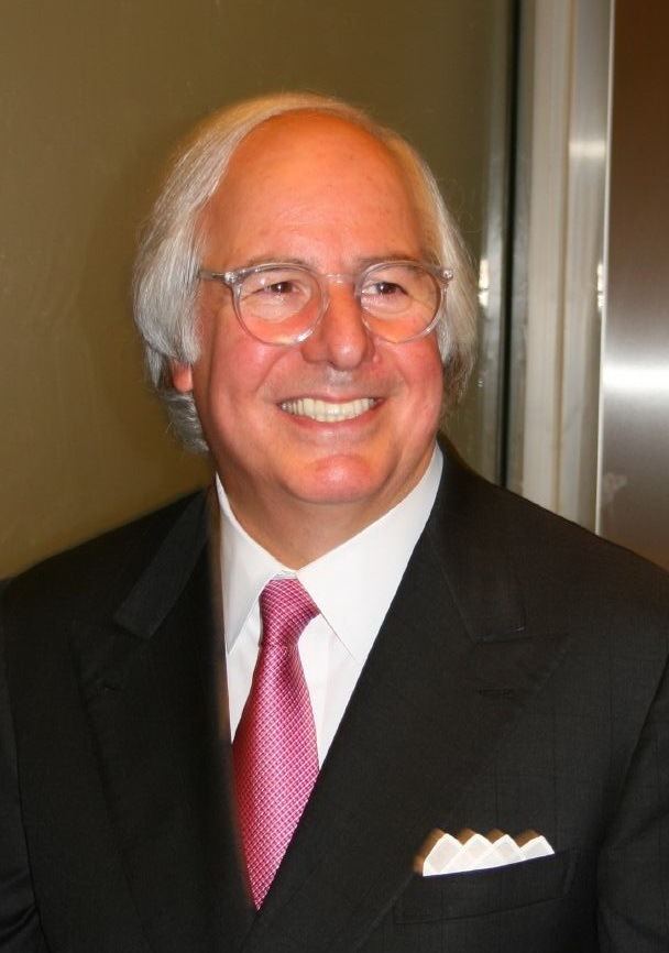 Frank Abagnale smiling and wearing a black coat over a white suit and a red necktie as well as an eyeglass.