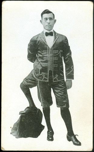 Frank Lentini's left foot standing on a backpack. Frank with a serious face is wearing shorts, high socks, shoes, and a long sleeve under a bow tie and coat