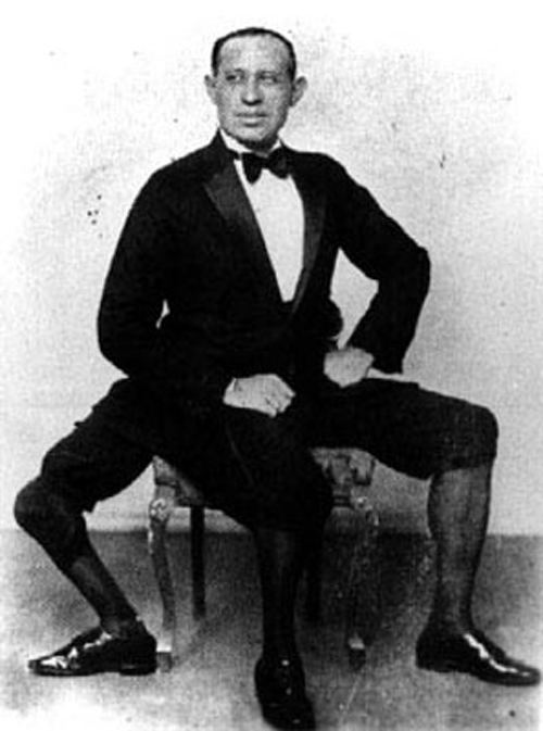 Frank Lentini sitting on the chair while looking afar with a serious face and wearing shorts, stockings, shoes, and a long sleeve under a bow tie and coat