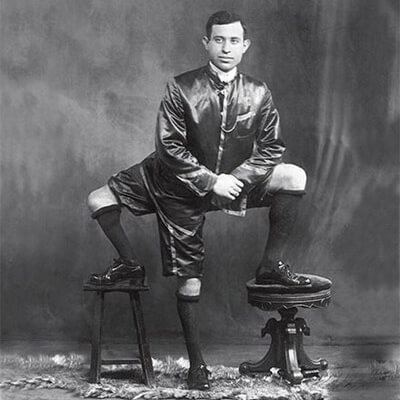 Frank Lentini posing while his left and right foot standing on the chair with a serious face, wearing shorts, shoes, high socks, and long sleeve shirt