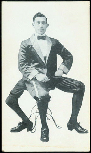 Frank Lentini with a tight-lipped smile while sitting on the chair and wearing shorts, high socks, shoes, and a long sleeve under a bow tie and coat