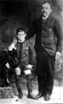 Young Frank Lentini holding the hand of the man beside him while his left foot is on the top of a rock. Frank with a serious face is wearing a long sleeve and boots while the man beside him, with a mustache, is wearing pants, shoes, and a long sleeve under a coat