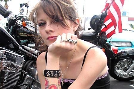 Frankie Abernathy hand on her chin and a motorcycle at her back while wearing a black sleeveless blouse, lip ring, black bracelet, and rings