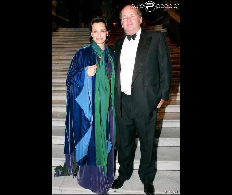 Kristin Scott Thomas smiling and wearing a blue dress and green scarf while François Olivennes wearing a coat, long sleeves, and bow tie