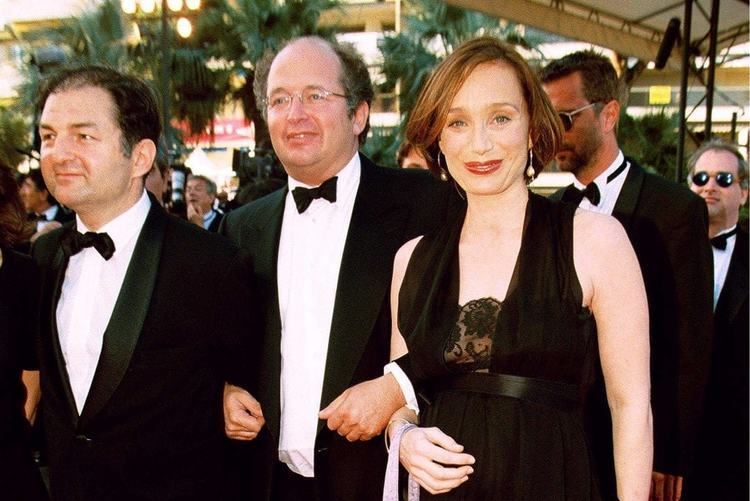 François Olivennes smiling and wearing a black coat, white long sleeves, and bow tie while Kristin Scott Thomas wearing a black dress