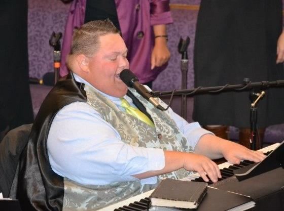 Freddie Combs wearing light blue long sleeves and black and gray vest while singing and playing piano