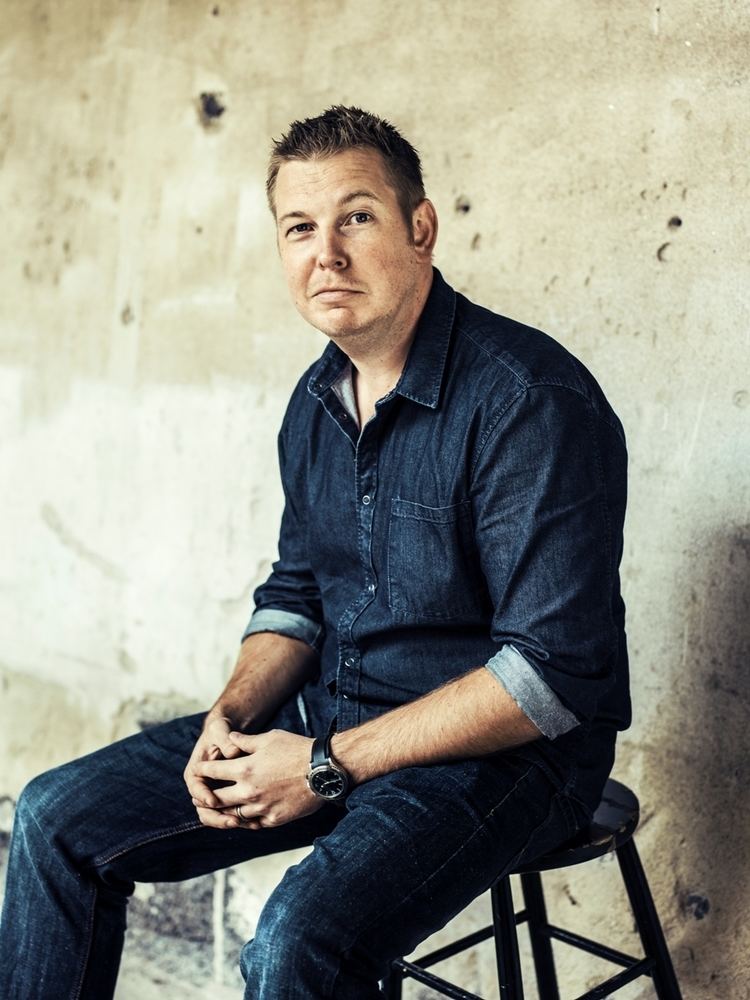 Fredrik Backman smiles while sitting on a chair wearing a watch, long sleeve, and jeans