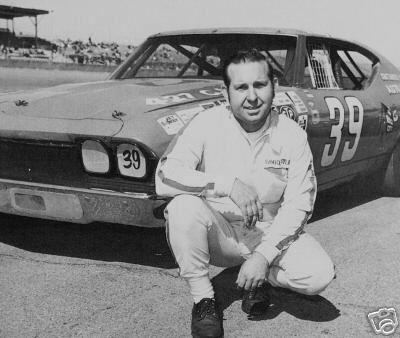 Friday Hassler is smiling in front of his racing car, wearing a white jacket, white pants, and black shoes.