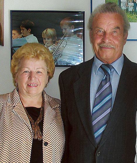 Josef Fritzl wearing a suit and a tie while Rosemarie Fritzl wearing earrings, a necklace, a striped blazer, and a brown shirt with blonde hair.
