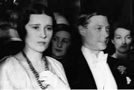 Thelma Furness and the Prince of Wales are seated next to each other, along with a group of people in the back. On the left is Thelma Furness, serious, sitting with black hair, wearing a white dress with white gloves and a necklace. Next to her is the Prince of Wales, smiling, sitting with black hair, wearing a white shirt with a bowtie and a black coat.