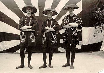 Three men were standing with their arms crossed at the back. There is a wall with a Japanese symbol of the rising sun and a plant on the right side. On the left, a man is serious, standing with arms crossed, wearing a traditional tube-sleeved Japanese coat with cross and Japanese letter patterns and a sandogasa hat. In the middle, a man is smiling, with arms crossed, wearing a traditional tube-sleeved Japanese coat with cross and Japanese letter patterns and a sandogasa hat. On the right, a man is serious, standing with arms crossed, wearing a traditional tube-sleeved Japanese coat with cross lines box with dot in the middle and Japanese letter patterns and a sandogasa hat.