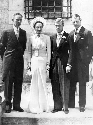 On the left, the man is serious, standing with both arms at his back. He has black hair, a white shirt and necktie, black pants, and a black coat. 2nd from left, Wallis Simpson, smiling, standing with both hands at her side, has black hair and is wearing a wedding dress with a hat. 3rd from left, Edward VIII is serious, standing with his hands down, has black hair, and is wearing a white polo with necktie, black shoes, black pants, and a jacket. On the right is a man standing behind Edward VIII. He has black hair and wears a white polo, a black necktie, a black coat and pants. On the left, the man is serious, standing with both arms at his back. He has black hair, a white shirt and necktie, black pants, and a black coat.
