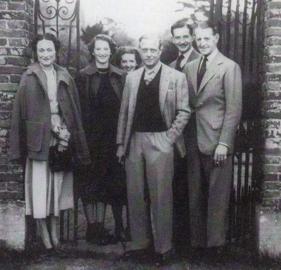 A large gate at their back, from the left, a woman is standing, her right hand holding her wrist and her left hand holding a black bag, has black hair, wearing a jacket, black shoes, and a white dress. 2nd from the left a woman is smiling, standing, has short black hair wearing a black dress black shoes and black coat, at her back is a girl smiling has short black hair, 4th from the left is a man smiling, standing, left hands on the pocket, has white hair, wearing a white polo, black shirt, white necktie, gray pants black shoes, and white coat, 5th from left is a man smiling, standing, has black hair, wearing polo black necktie and black coat, at the right is Fruity Metcalfe, smiling while standing, wearing a gray coat, white polo with black tie and black shoes.