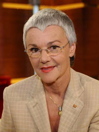 Gabriele Krone-Schmalz is smiling having gray hair with pointed bangs in a dim-lit background while wearing a gold pendant necklace, and a patterned-beige coat with a gold brooch.