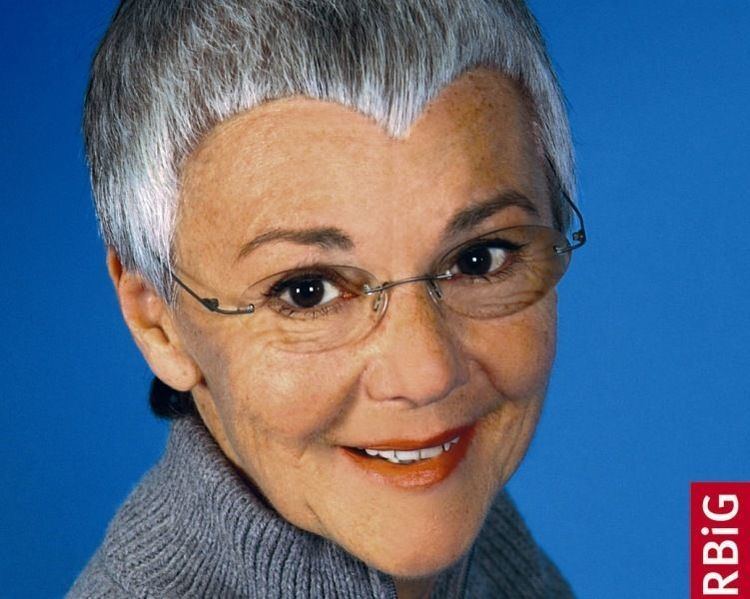 Gabriele Krone-Schmalz is smiling and has gray hair with pointed bangs in a blue background. Gabriele is wearing eyeglasses, orange lipstick, a gray knitted turtle neck blouse.