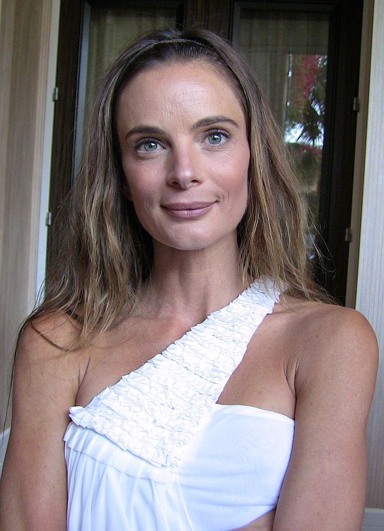 Gabrielle Anwar smiling with a glass door on her background, she has blonde hair down wearing a one-shoulder white top
