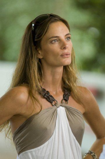 Gabrielle Anwar looks serious while looking at someone with both hands on her waist, has a blonde hair, wearing a bracelet on her left hand and a braless sexy white and gray top