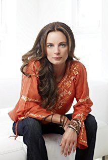 Gabrielle Anwar looks serious while sitting on a white couch while both hands crossed together, she has her black hair down wearing black bracelets and an orange long sleeve top and jeans