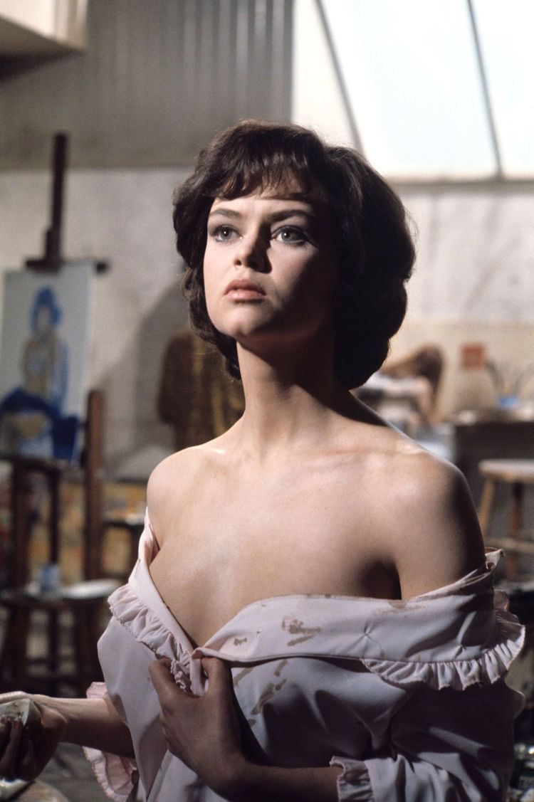 Gabrielle Drake looking above, with wavy short hair, and wearing a sexy blouse showing almost her breast.